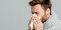 Caucasian man with beard wearing gray scarf blowing nose into tissue with pained expression against neutral background, depicting cold or flu symptoms.