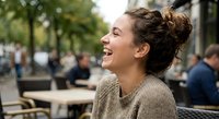 Young woman laughing joyfully at outdoor cafe terrace with friends in background. Genuine happiness and social connection in urban setting.