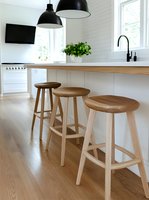 Modern minimalist kitchen interior with wooden bar stools at white counter, black pendant lamps, and natural light from windows creating bright scandinavian atmosphere.