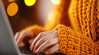 Female hands typing on laptop keyboard wearing cozy orange knit sweater with soft bokeh lights in background, creating warm autumn mood for remote work.