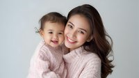 Young Asian mother and baby daughter in matching pink knit sweaters share tender moment against light background, expressing joy and genuine connection.