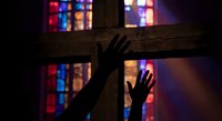 Silhouette of hands raised in prayer against wooden cross with colorful stained glass window light creating spiritual atmosphere for worship and faith.
