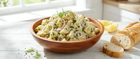 Creamy potato salad with fresh herbs and vegetables served in wooden bowl with sliced bread on white table for lunch or dinner meal preparation.