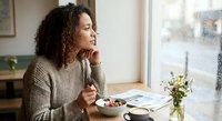 Middle-aged woman with curly hair enjoying healthy breakfast at home, looking thoughtfully out window while eating yogurt with berries in cozy kitchen setting.