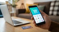 Person holding smartphone with shopping cart app interface for online retail purchase. Laptop and credit card on wooden desk in modern home office setting.