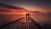 Couple standing on wooden pier at sunset, holding hands while watching dramatic orange and red sky over calm ocean water, romantic travel destination for honeymoon or anniversary celebration.