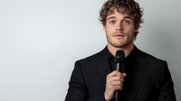 Young Caucasian male with curly hair wearing black suit holding microphone against light background, professional headshot portrait for business presentation or public speaking.