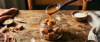 Wooden spoon drizzling golden caramel sauce into a rustic glass jar filled with soft caramel candies on a vintage wooden table.