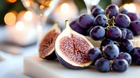 Fresh ripe purple figs cut in half showing juicy red flesh alongside dark grape cluster on white surface with soft bokeh lights in background.