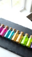 Colorful liquid-filled glass vials with cork stoppers arranged on dark slate surface near bright window, featuring vibrant magenta, cyan, yellow, and lime green hues for wellness, aromatherapy, or potion design projects.