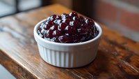 Fresh homemade cranberry sauce with whole berries in white ceramic ramekin on rustic wooden table, selective focus showing glossy texture and deep red color.