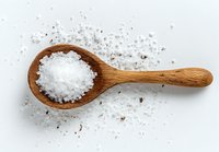 Coarse sea salt crystals on wooden spoon against white background, scattered salt grains around for culinary and food photography.