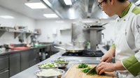 Professional male chef with glasses preparing fresh green vegetables on cutting board in modern commercial restaurant kitchen during food preparation service.
