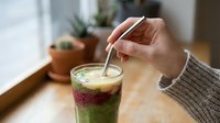 Young woman stirring layered matcha and berry smoothie drink with metal straw in glass, indoor setting with potted plants and natural light for beverage and healthy lifestyle content.