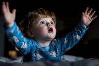 Adorable toddler boy in blue pajamas reaching up with wonder and excitement, expressing joy and amazement with outstretched arms against dark background.