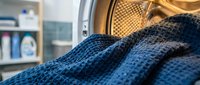 Blue knitted sweater being loaded into modern washing machine drum in bright laundry room with detergent bottles on shelves in background.