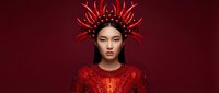 Young woman with East Asian features wearing elaborate red sequined garment and ornate spiky crown headdress against deep burgundy background, suitable for fantasy, fashion, editorial and creative design projects.