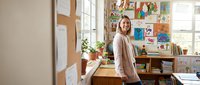 Smiling Caucasian female teacher standing in a colorful elementary classroom with children artwork on walls, bright and welcoming school environment.