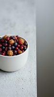 Mixed berries including blueberries, raspberries, and cloudberries in white bowl on concrete surface with neutral background for food styling and culinary projects.