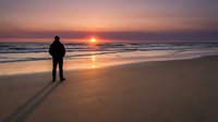 Man in dark jacket standing on sandy beach watching golden sunset over ocean waves, contemplative coastal scene for travel and lifestyle use.