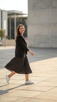Young woman with long brown hair, Caucasian ethnicity, wearing black blazer and polka dot midi skirt with white sneakers, walking in modern urban plaza with contemporary architecture.