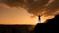 Silhouette of person with raised arms celebrating success on mountain cliff edge against dramatic orange sunset sky with clouds over canyon landscape.