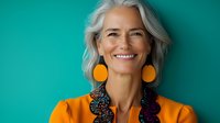 Confident mature woman with silver hair wearing vibrant orange outfit and statement earrings smiling against turquoise background. Modern aging and fashion concept.