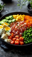 Fresh protein bowl with sliced prosciutto, boiled eggs, cucumber, mango, carrots, peas, and tomatoes arranged in colorful sections on dark plate. Food photography.