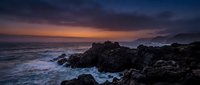 Dramatic rocky coastline at twilight with golden sunset sky reflecting on turbulent ocean waves and dark stone formations, suitable for travel and nature backgrounds.
