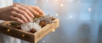 Hands reaching for natural spa items in wooden box with sea salt, star anise and dried herbs against soft bokeh background with floating light particles.