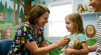 Female pediatric healthcare professional examining young Caucasian girl patient with stethoscope in colorful children's medical office with toys and decorations.