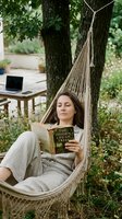 Middle-aged woman relaxing in hammock between trees, reading book in garden setting with laptop nearby, ideal for wellness and remote work lifestyle content.