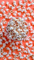 Fresh popcorn in white bowl on vibrant coral background, scattered kernels creating festive texture for movie night or party snack concept.