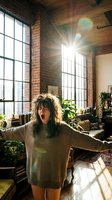 Young woman with dark wavy hair, Caucasian ethnicity, wearing oversized beige sweater, expressing joy with arms outstretched in sunlit loft apartment with exposed brick walls, large industrial windows, wooden floors, and potted plants.