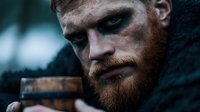 Intense close-up portrait of bearded redhead man with dramatic dark makeup and fierce expression against moody background, holding wooden mug in medieval style setting.
