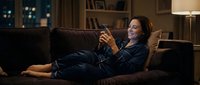 Woman relaxing on brown sofa at night, wearing dark shirt, holding smartphone, enjoying leisure time at home with warm lighting and shelves.