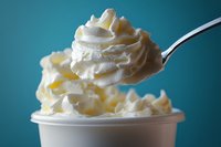 Fresh whipped cream lifted by spoon above white container, showing light, airy texture against turquoise background, macro food photography.