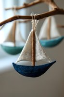 Handcrafted sailboat ornament with white canvas sail and navy blue hull hanging from natural branch, creating whimsical nautical decor against soft blurred background.