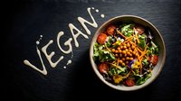 Fresh vegan salad bowl with chickpeas, greens, tomatoes, and edible flowers on dark slate with vegan text written in sauce.