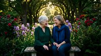 Senior woman and middle-aged woman sitting together on bench in blooming garden with purple and red flowers, enjoying conversation under tree canopy during golden hour.