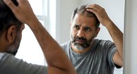 Middle-aged Hispanic man examining hair loss in bathroom mirror, touching scalp with concerned expression for male pattern baldness awareness campaigns.