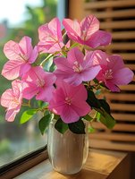 Vibrant pink bougainvillea flowers arranged in a clear glass vase on a wooden surface near a window, showcasing delicate petals and green foliage for home decor and floral arrangement inspiration.