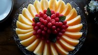Fresh cantaloupe slices arranged in sunburst pattern on glass platter with bright red raspberries and mint leaves in center, elegant fruit platter for entertaining.