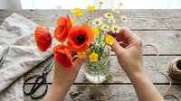 Hands arranging vibrant red poppies and yellow daisies in glass vase on wooden table with scissors and linen cloth, perfect for floral design and home decor projects.