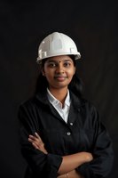 Young South Asian female engineer wearing white hard hat and black workwear stands confidently with crossed arms against dark background, representing diversity in STEM fields.