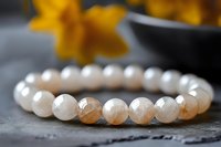 Elegant natural moonstone beaded bracelet with round gemstones on dark stone surface, yellow flowers in background creating soft bokeh effect.