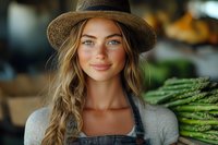 Young Caucasian woman farmer in straw hat and denim overalls holding fresh asparagus at local market, with natural smile and braided blonde hair in soft lighting.