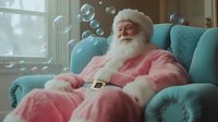 Senior man with white beard and rosy cheeks wearing pink fuzzy suit sitting in turquoise armchair surrounded by floating soap bubbles near window for holiday entertainment.