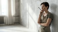 Young man with dark hair and East Asian ethnicity wearing casual olive green shirt and work apron, holding pencil thoughtfully against chin while leaning on white wall in bright studio space, contemplating design or construction project.