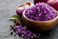 Purple sea salt crystals in wooden bowl with spices and herbs on dark stone background, macro food photography for culinary design and packaging.